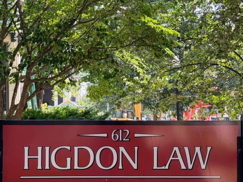 A red street sign reading 612 Higdon Law positioned in front of green trees.